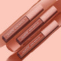 Three ColourPop Ultra Matte Lip tubes in the Warmed Up bundle rest diagonally atop wide nude‑brown swatches, showcasing a full‑coverage ultra matte finish with clear barrels and blush pink star caps on a peach backdrop.