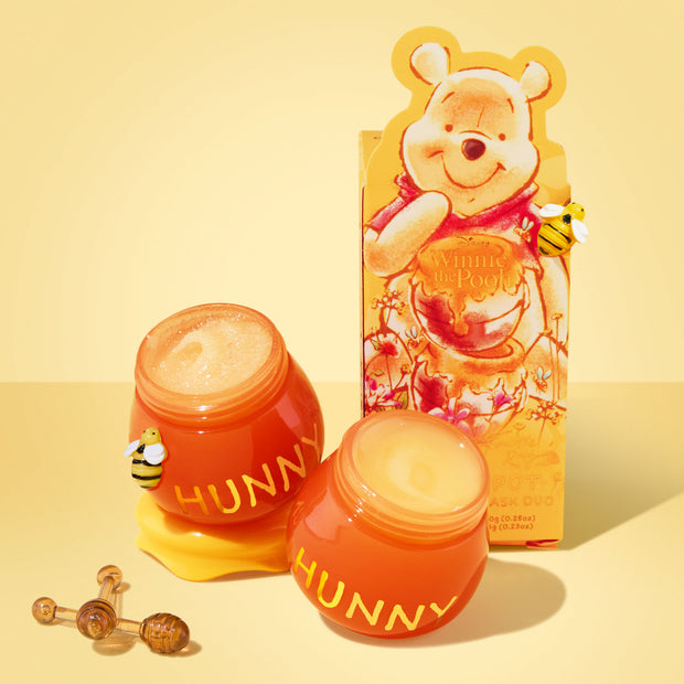 ColourPop Hunny Pot Lip Care Kit shown open, two honey-pot jars labeled HUNNY reveal lip scrub and lip mask with honey-like texture, bees and honey dipper accents, set before Winnie the Pooh–themed packaging on a warm yellow background.