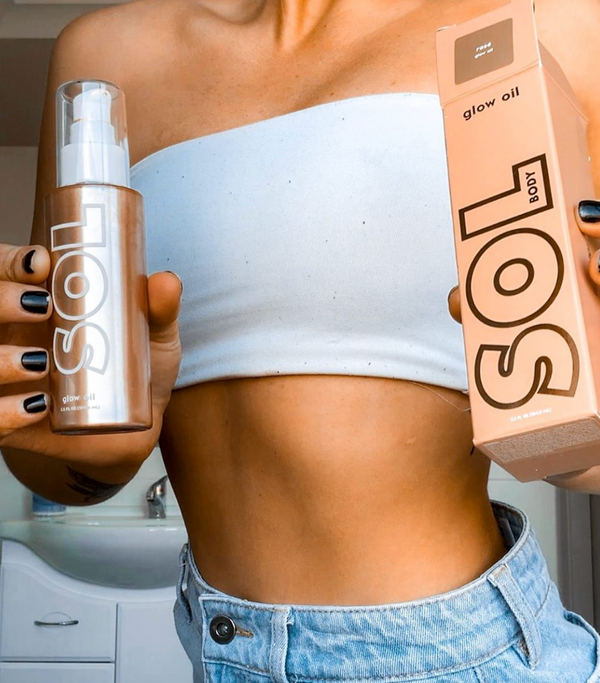 ColourPop SOL Glow Oil Rosè bottle and box held by a person, highlighting the bronze shimmery body oil pump and peach packaging; shown at torso level in a bright bathroom, with a white bandeau and denim shorts.