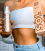 ColourPop SOL Glow Oil Rosè bottle and box held by a person, highlighting the bronze shimmery body oil pump and peach packaging; shown at torso level in a bright bathroom, with a white bandeau and denim shorts.