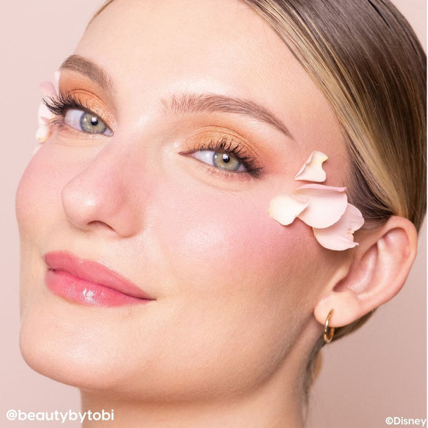 Warm gold shimmer eyeshadow illuminating lids with a softly defined crease and long lashes, shown in a close-up with pink rose petals on the cheek and glossy lips, highlighting the Beauty and the Beast ColourPop Shadow Palette’s romantic tones.
