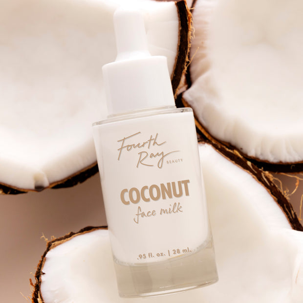 Fourth Ray Beauty Coconut Face Milk dropper bottle tilts upright, showing .95 fl oz/28 mL label, nestled among halved coconuts on a soft beige background; hydrating ColourPop Face Milk skincare with a creamy white formula.