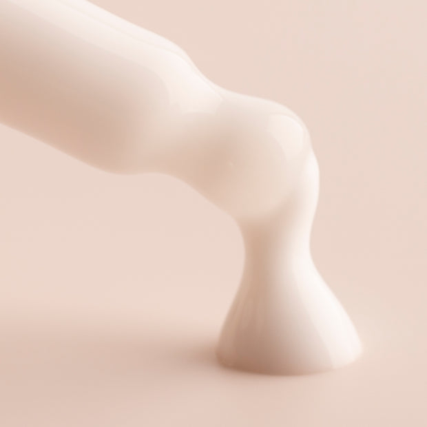 Coconut Face Milk, a ColourPop Face Milk, pours in a glossy white ribbon and pools on a soft beige surface, close-up emphasizing its lightweight, hydrating texture.