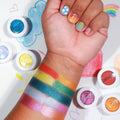 Pug Life Super Shock Shadow swatched on a forearm, showing smooth, metallic payoff, surrounded by open ColourPop Super Shock Shadow pots in rainbow hues; hand with rainbow nail art rests over a doodled cloud and rainbow background.