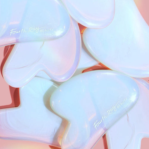 Opalite Crystal Gua Sha tools stacked and gleaming with an iridescent finish on a pink backdrop, Fourth Ray Beauty branding visible, showcasing a facial massage scraper from ColourPop Skincare Tools And Accessories for contouring and de-puffing.