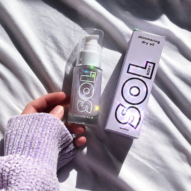 SOL Body Opaltini shimmering dry oil bottle with pump - sparkling with iridescent glitter - held in a hand beside its holographic SOL box on sunlit white bedding; ColourPop SOL Shimmering Dry Oil for a luminous body glow.