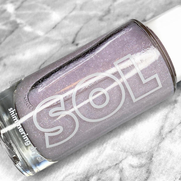 ColourPop SOL Shimmering Dry Oil Opaltini bottle glinting with iridescent lavender shimmer, lying on a gray marble surface; clear glass with bold SOL logo and white cap shows the pearly body-glow dry oil inside.