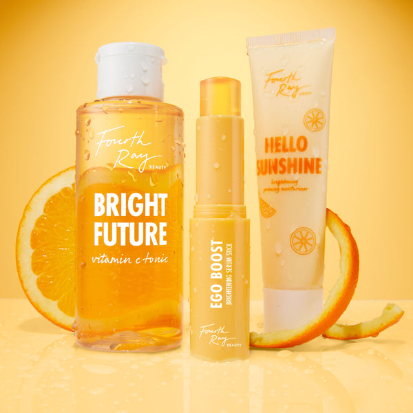 Bright Future Vitamin C Tonic bottle from Fourth Ray Beauty, beaded with water, stands between Ego Boost Brightening Serum Stick and Hello Sunshine moisturizer on a glossy orange surface with citrus slices; ColourPop skincare.