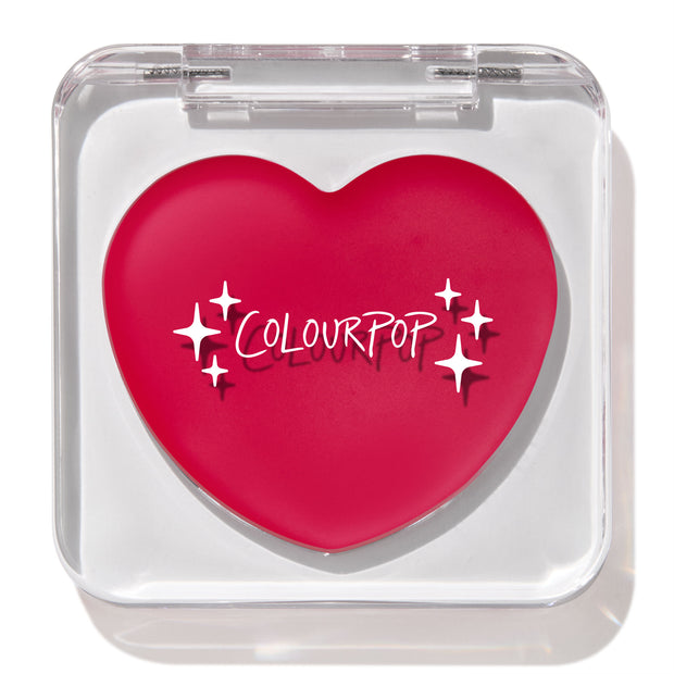 Heart-shaped hot pink compact with white “ColourPop” logo and sparkles rests closed in a clear square case, featuring the ColourPop Lip and Cheek Balm in Girls Night, a rosy multitasking balm for lips and cheeks.