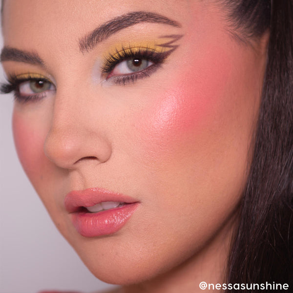ElectroBall ColourPop Cream Blush delivering a dewy, vibrant coral‑pink flush on the cheek, showcased in a close-up with bold yellow winged eyeliner, long lashes, and glossy pink lips for a fresh, luminous finish.