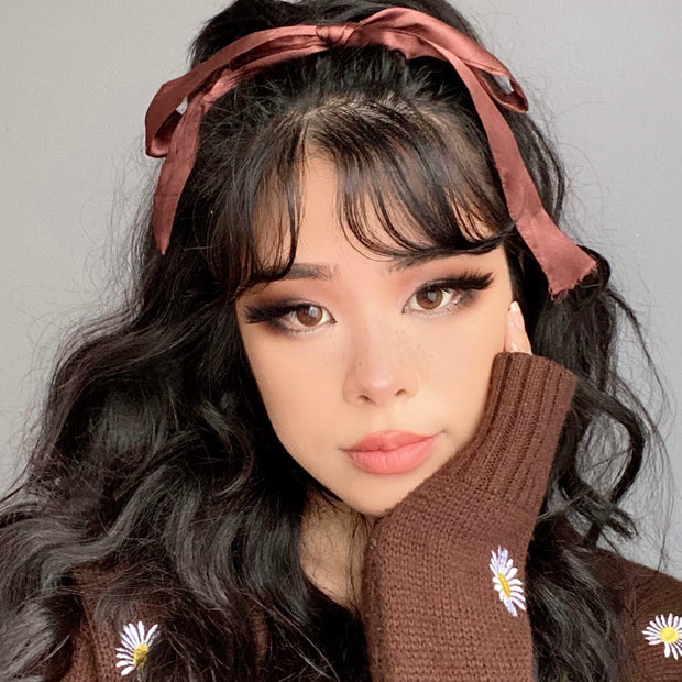 Model showcases a soft neutral eye using the ColourPop Gone Matte shadow palette, blending taupe-brown mattes with a smoky wing; wavy dark hair tied with a brown ribbon and a brown sweater with daisy embroidery in a studio close-up.