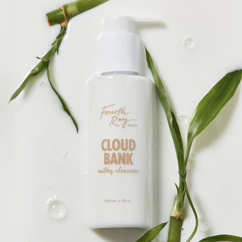 Cloud Bank Milky Cleanser bottle by Fourth Ray Beauty rests on a glossy, milk-like surface with water droplets, framed by green bamboo leaves; white pump-top 5.8 fl oz (171 ml) packaging, presented as a gentle ColourPop Face Cleanser.