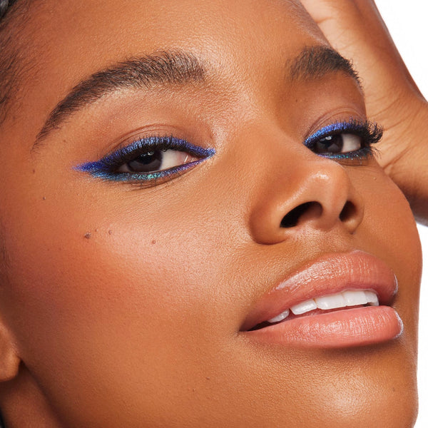 Ventura Blvd ColourPop Chrome Crème Gel Liner sweeping a bold cobalt-blue wing with a teal duochrome shift along upper and lower lash lines, shown in a close-up on a model with smooth skin and glossy nude lips against a bright studio backdrop.