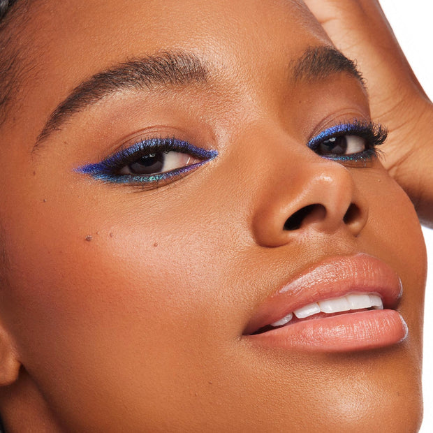 Ventura Blvd ColourPop Chrome Crème Gel Liner sweeping a bold cobalt-blue wing with a teal duochrome shift along upper and lower lash lines, shown in a close-up on a model with smooth skin and glossy nude lips against a bright studio backdrop.