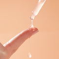 Dropper dispensing milky Reveal 10% AHA Serum onto a fingertip, a glossy bead stretching and dripping against a soft peach background, showcasing the exfoliating ColourPop Face Serum for smooth, radiant skin.