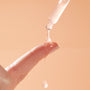Dropper dispensing milky Reveal 10% AHA Serum onto a fingertip, a glossy bead stretching and dripping against a soft peach background, showcasing the exfoliating ColourPop Face Serum for smooth, radiant skin.