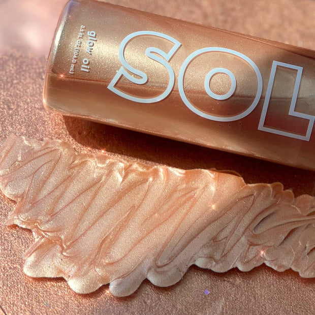 Rosè SOL Glow Oil bottle resting on a rose-gold surface, with a zigzag swatch of pearly rose shimmer showing its luminous finish; ColourPop SOL Glow Oil body highlighter in shade Rosè for a sun-kissed glow.