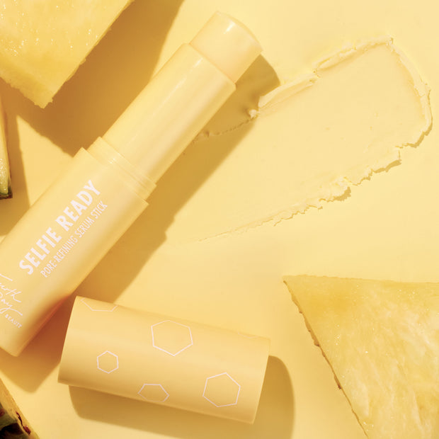 Selfie Ready pore-refining ColourPop Serum Stick resting open with its cap beside it and a creamy swipe, on a monochromatic yellow backdrop styled with pineapple slices, showcasing the portable serum stick’s smooth texture and bright vibe.