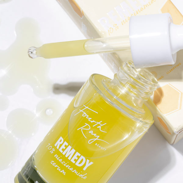 Fourth Ray Beauty Remedy + Rainfall ColourPop Face Serum drips from a white dropper into pale yellow pools, showing a 10% niacinamide formula in a clear glass bottle beside its cream carton on a white surface.
