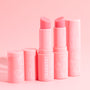 Strawberry lip balm sticks stand uncapped, revealing rosy bullets, in glossy pink Fourth Ray Beauty tubes with white strawberry icons on a soft pink backdrop - Strawberry flavor ColourPop Lip Balm for soft, hydrated lips.