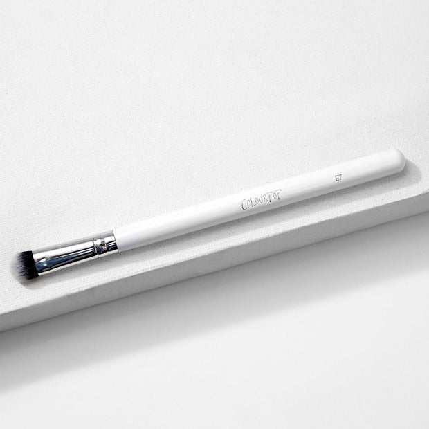 ColourPop Medium Dome Brush blending detail eye makeup - white-handled makeup brush with chrome ferrule and domed black‑to‑white bristles, marked E7 - resting diagonally on a minimalist white textured backdrop.