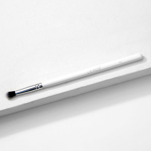 ColourPop Small Tapered Brush resting diagonally on a white textured platform, featuring a slim white handle, chrome ferrule, and small tapered black-and-white bristles for precise eyeshadow blending and detail work.