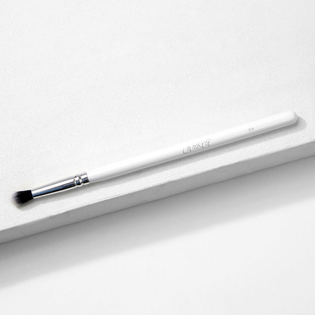 ColourPop Small Tapered Brush resting diagonally on a white textured platform, featuring a slim white handle, chrome ferrule, and small tapered black-and-white bristles for precise eyeshadow blending and detail work.