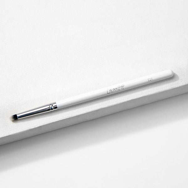 ColourPop Small Pencil Brush makeup brush lying diagonally on a white textured block, showing a slim white handle, silver ferrule, and small tapered bristles for precise eyeshadow application and smudging.