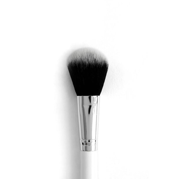 Blush brush with tapered, fluffy black-to-white synthetic bristles blending powder across cheeks, anchored by a chrome ferrule and white handle on a clean white background; ColourPop Makeup Brush for precise, soft application.