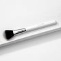 Blush Brush - ColourPop Makeup Brush with soft black-to-white bristles and chrome ferrule - lying diagonally on a white textured platform, white handle stamped ColourPop, ideal for sweeping and blending powder blush.