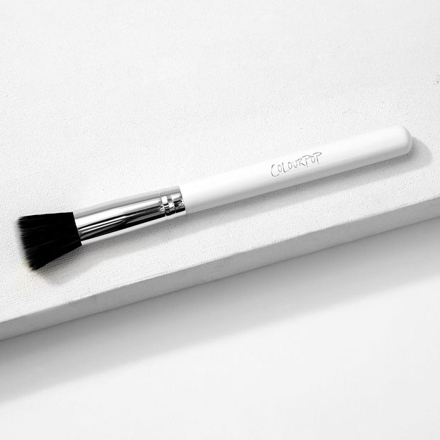 ColourPop stippling makeup brush resting diagonally on a white textured block, featuring duo-fiber black-and-white bristles, a shiny silver ferrule, and a white handle with the ColourPop logo.