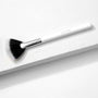 ColourPop Fan Brush makeup brush resting diagonally on a white textured block, featuring duo-fiber black-to-white fanned bristles, a silver ferrule, and a white handle embossed with ColourPop, designed for sweeping highlighter.