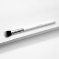 ColourPop Small Fluff Brush lies diagonally on a white textured block, showing soft synthetic black-to-white bristles, a chrome ferrule, and a white handle; a versatile makeup brush for precise blending in a clean studio setting.