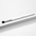ColourPop Blending Brush makeup brush resting diagonally on a white textured surface, featuring fluffy angled black-to-white bristles, a silver ferrule, and a slim white handle, made for seamless eyeshadow blending.