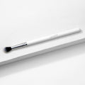 ColourPop Tapered Blending Brush resting diagonally on a white textured surface, featuring a white handle, silver ferrule, and soft tapered black-and-white bristles for seamless eyeshadow blending and crease definition.