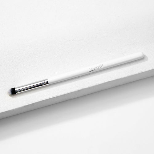 ColourPop Pencil Brush resting diagonally on a white textured block, a makeup brush with a slim white handle and logo, silver ferrule, and small tapered black-and-white bristles for precise eyeshadow and eyeliner smudging.
