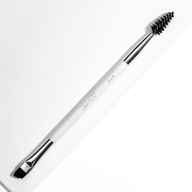ColourPop Brow Dual Ended makeup brush resting diagonally, featuring an angled flat brow brush for filling and a black spoolie for grooming, white handle with chrome ferrules, on a bright white textured background.