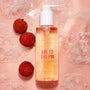 Clear pump bottle of Fourth Ray Beauty AM to the PM Gel Cleanser by ColourPop glistening with golden gel, sitting amid foamy suds on a pink surface next to lychees; 5.8 fl oz face cleanser.