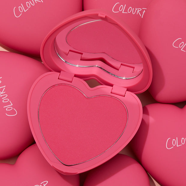 ColourPop Pressed Powder Blush Text Me opens to reveal a heart-shaped vivid pink pan with built-in mirror, set in a matte hot‑pink compact and surrounded by matching ColourPop heart cases, highlighting a romantic, travel-friendly cheek color.