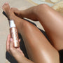 ColourPop SOL Glow Oil Rosè bottle with pump held beside sunlit legs, the shimmering body oil leaving a rosy bronze glow on skin against a stone patio backdrop.