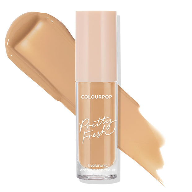 ColourPop Pretty Fresh Creamy Concealer in Medium 100 W stands upright in a clear tube with a blush-pink cap, showing a warm medium beige shade as two smooth swatches sweep behind on a white background, highlighting its hyaluronic creamy formula.