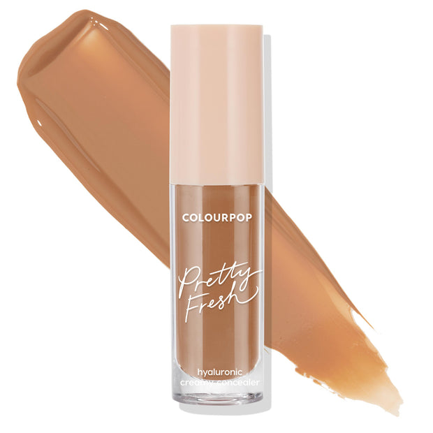 ColourPop Pretty Fresh Creamy Concealer in Dark 158 W stands upright with a blush-beige cap and clear tube marked “Pretty Fresh” and “hyaluronic,” while a warm tan shade swatch streaks behind on a white background.