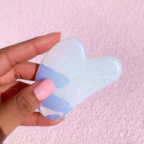 Opalite Crystal Gua Sha facial massage tool held in a hand, showing its iridescent blue‑white stone and curved edges, branded Fourth Ray Beauty, against a pink textured background; part of ColourPop Skincare Tools And Accessories.