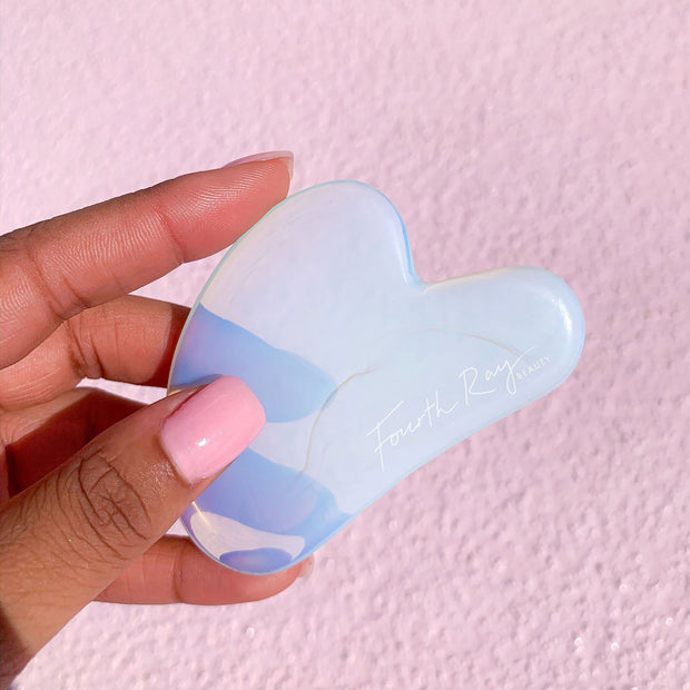 Opalite Crystal Gua Sha facial massage tool held in a hand, showing its iridescent blue‑white stone and curved edges, branded Fourth Ray Beauty, against a pink textured background; part of ColourPop Skincare Tools And Accessories.