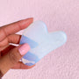 Opalite Crystal Gua Sha facial massage tool held in a hand, showing its iridescent blue‑white stone and curved edges, branded Fourth Ray Beauty, against a pink textured background; part of ColourPop Skincare Tools And Accessories.