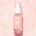 ColourPop SOL Shimmering Dry Oil in Soft Pink stands upright in a clear bottle with white pump and cap, fine shimmer suspended, SOL logo visible, catching light on a sparkly blush-pink background.