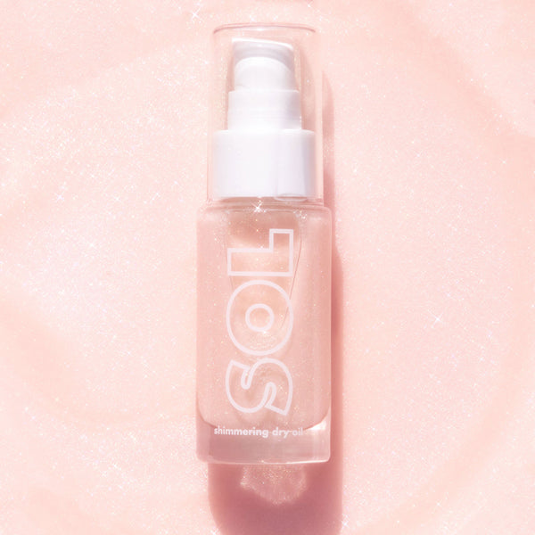 ColourPop SOL Shimmering Dry Oil in Soft Pink stands upright in a clear bottle with white pump and cap, fine shimmer suspended, SOL logo visible, catching light on a sparkly blush-pink background.