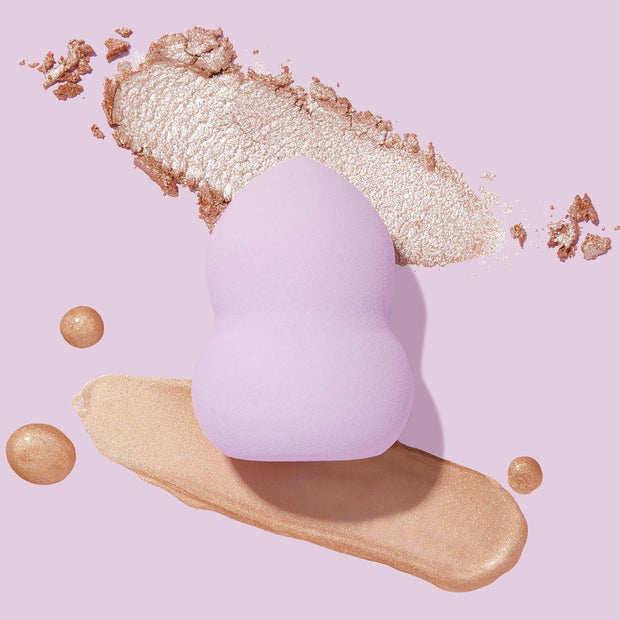 Hourglass Blending Sponge blending shimmery gold highlighter, a lavender, double-curved makeup sponge set on a pastel purple backdrop with liquid and crushed powder swatches, ideal for ColourPop Makeup Tools & Accessories.