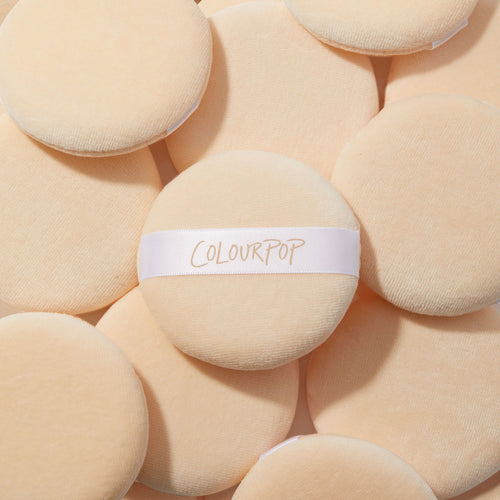 ColourPop Powder Puff showing a soft beige velour round with a white elastic hand strap labeled ColourPop, lying atop a pile of matching puffs - plush tool for applying and setting powder, part of ColourPop Makeup Tools & Accessories.