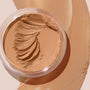 ColourPop SOL Face & Body Bronzing Balm in shade Light sits in a clear round jar with creamy swirls, with warm golden-tan balm swatched around it on a beige surface, showcasing a smooth, blendable texture and natural dewy finish.